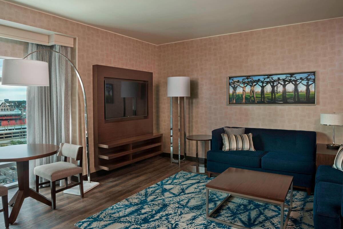 A hotel room with a wooden floor, a blue sofa, a TV mounted on a wooden panel, a small round table with chairs, a patterned rug, and a painting on the wall.