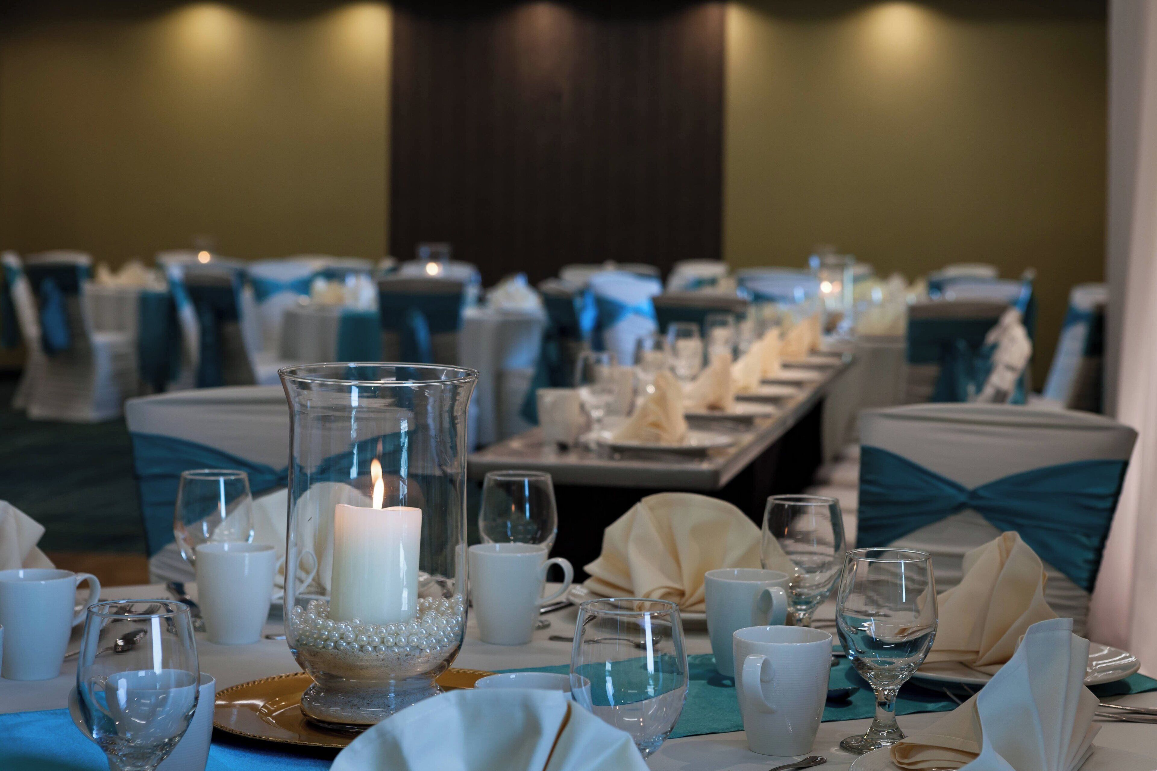 A banquet table set for a formal event with a lit candle centerpiece, white tableware, and teal chair covers.