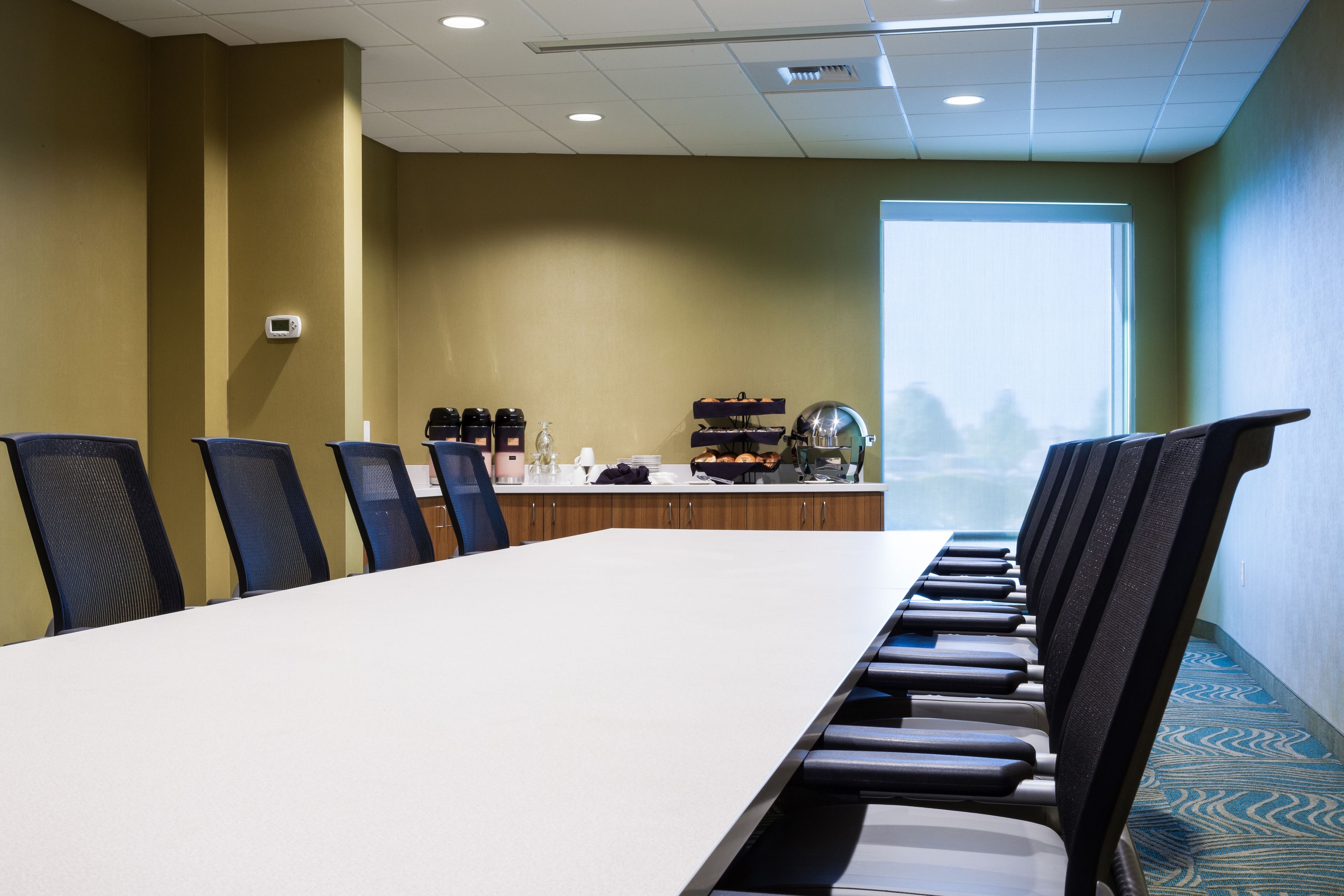 A conference room with a long table, chairs, a coffee station, and a view through a large window.