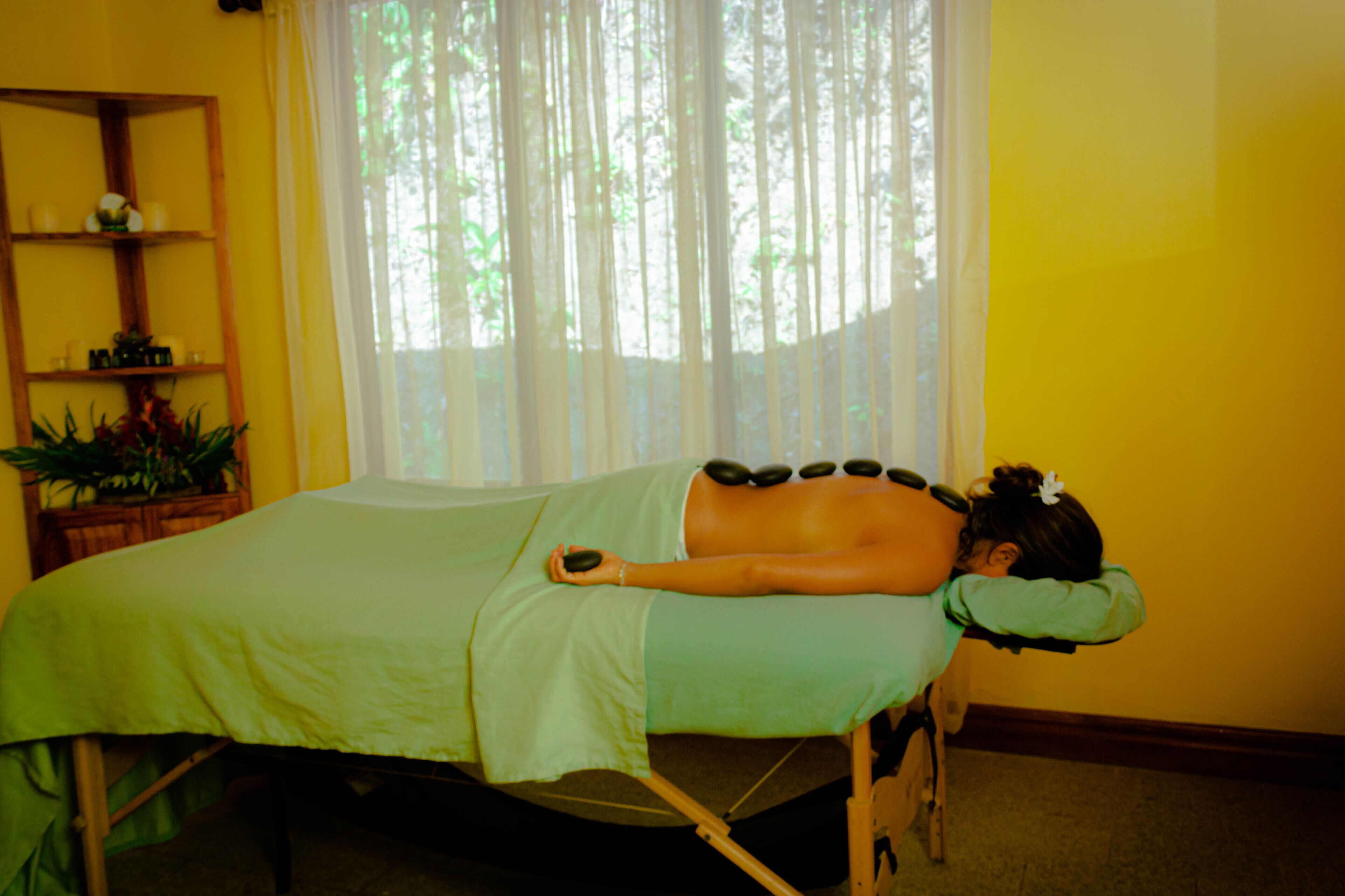 Une salle de massage équipée d’un lit, d’une étagère en bois où sont rangés divers objets, et d’une fenêtre avec des rideaux.