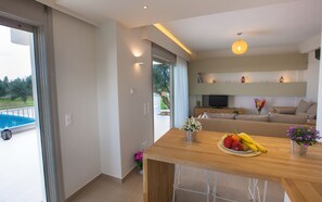 A modern living room with a wooden dining table, a sofa, and a TV. There is a balcony with a pool and a garden visible through the glass doors.