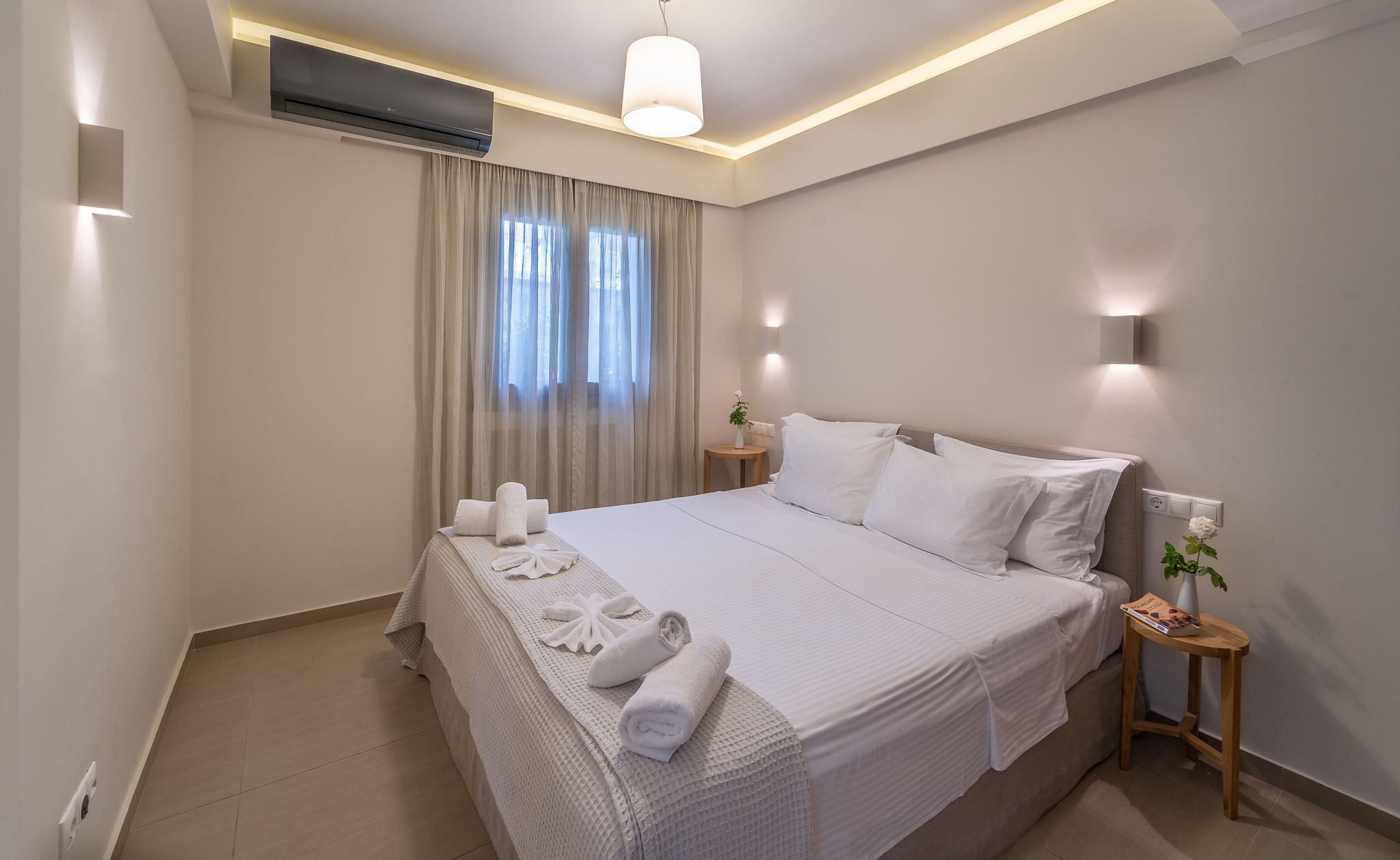 Ein modernes Hotelzimmer mit einem großen Bett, einem Nachttisch mit einem Buch, einer kleinen Pflanze und einer Wand-Klimaanlage.
