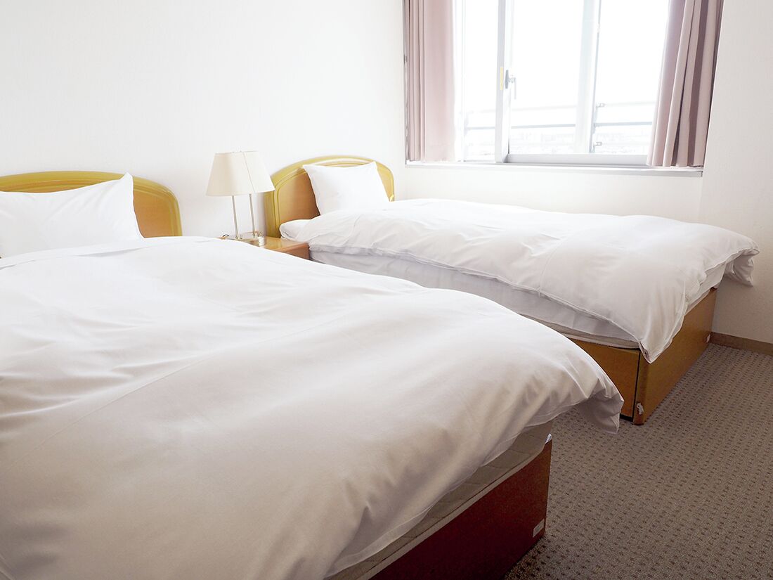 Quarto de hotel com duas camas, uma mesinha de cabeceira com luminária e uma janela com cortinas.