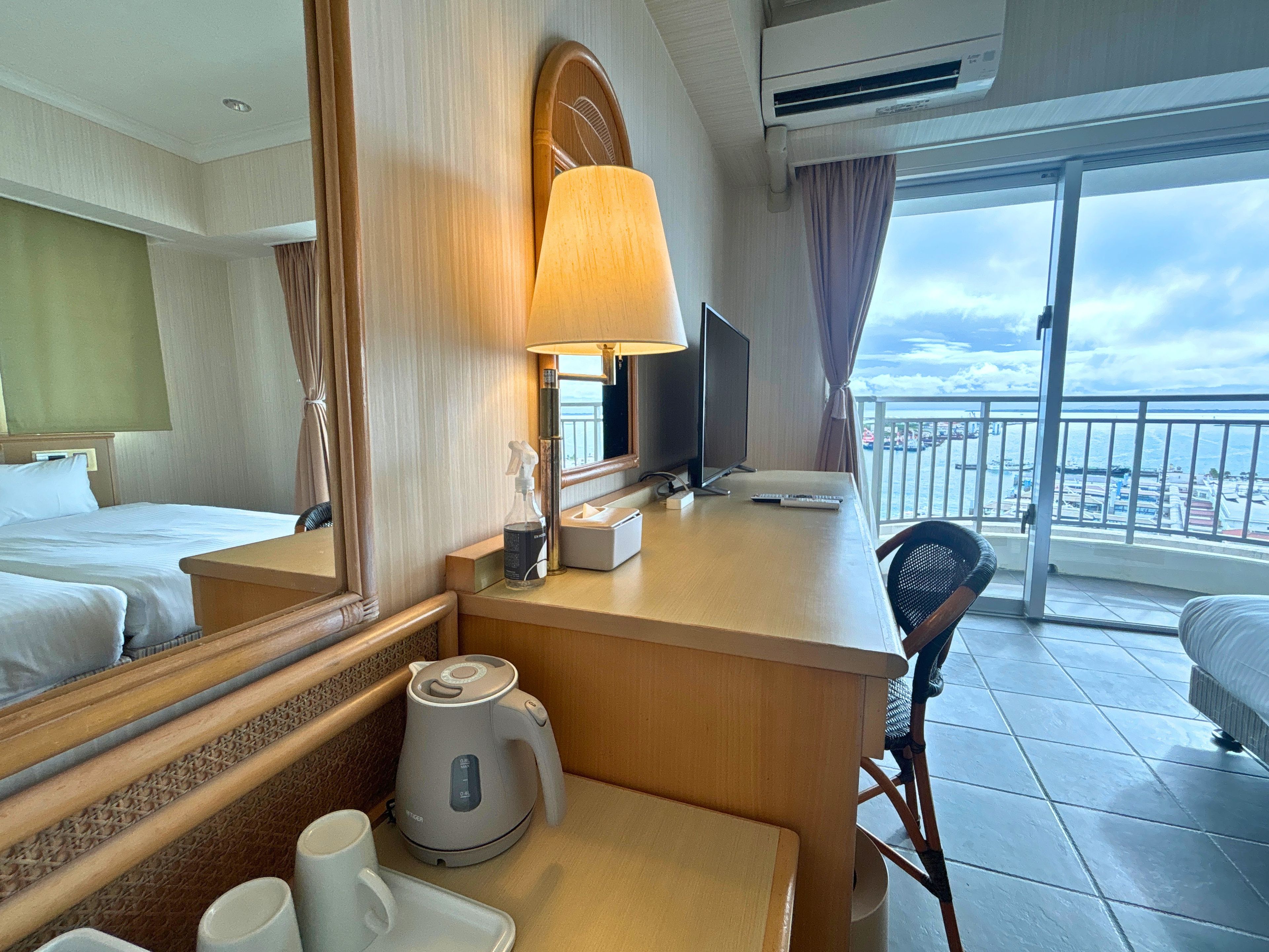 Una habitación de hotel con cama, escritorio con lámpara, televisor, cafetera y vista a la ciudad por la ventana.