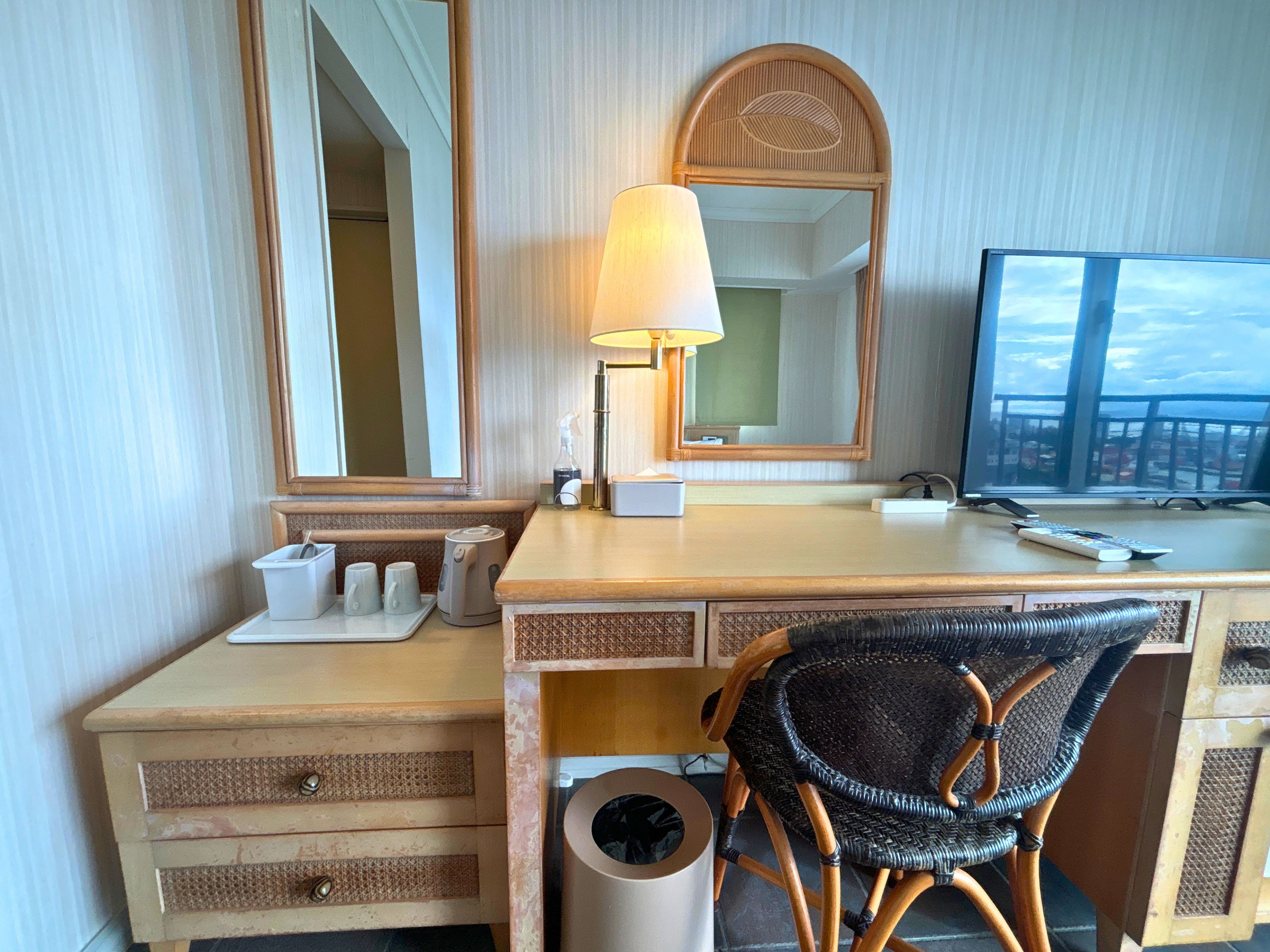 A hotel room with a wooden desk, a chair, a mirror, a lamp, a television, and a window with a view.