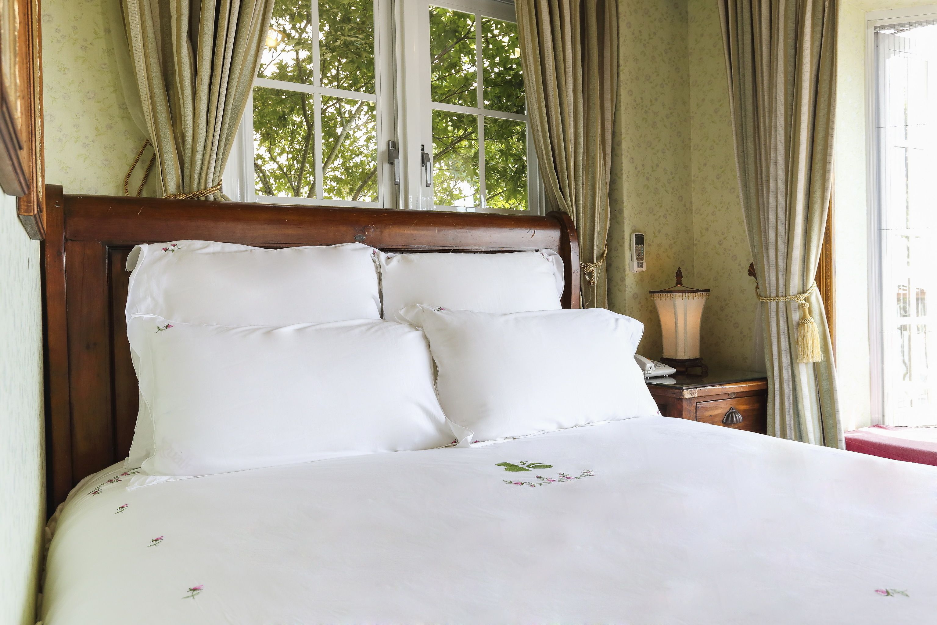 A neatly made bed with white linens and floral patterns, a wooden headboard, and a window with curtains.