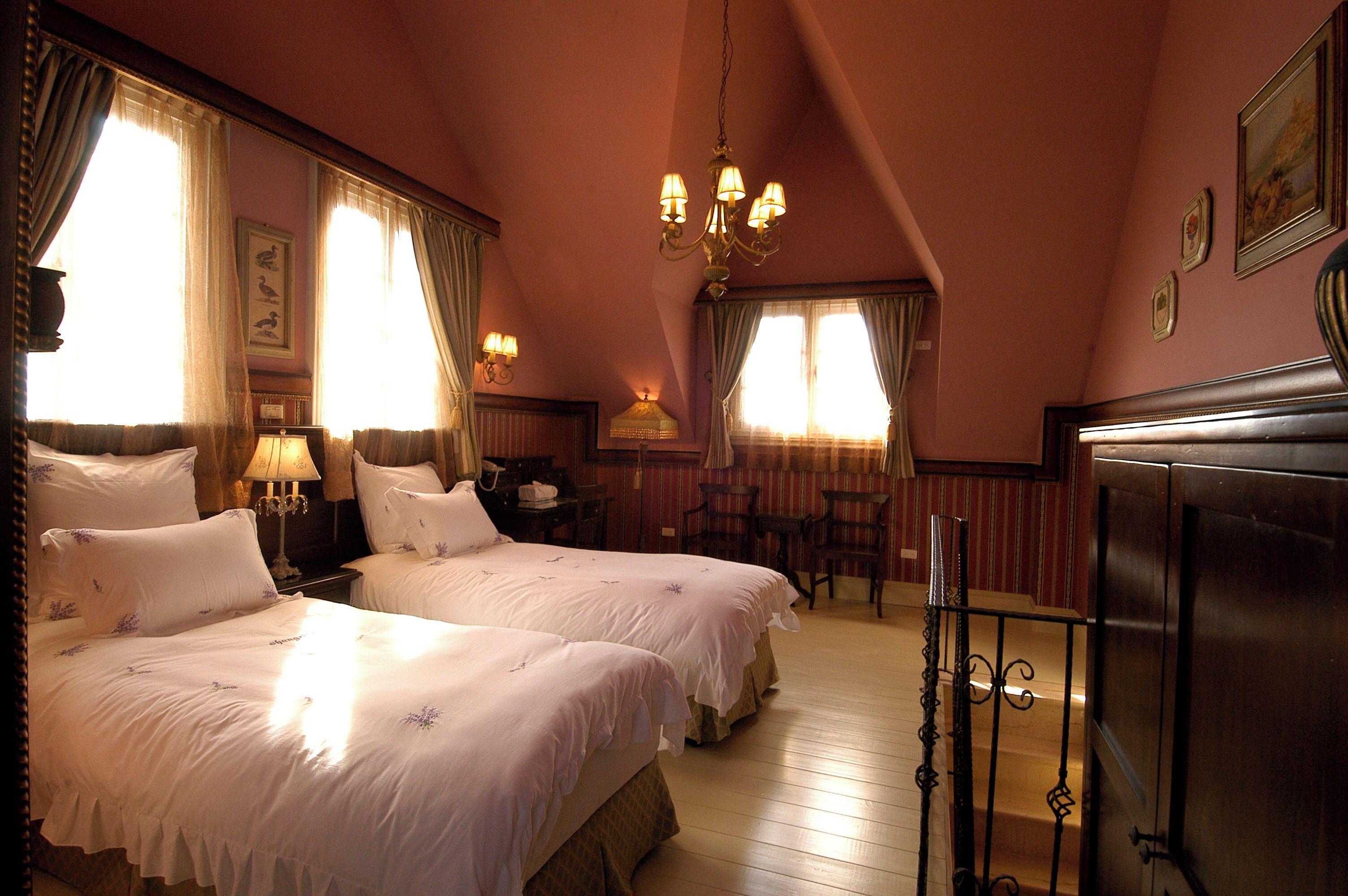 A hotel room with two beds, a chandelier, a desk with a lamp, and a view of the outside through the windows.