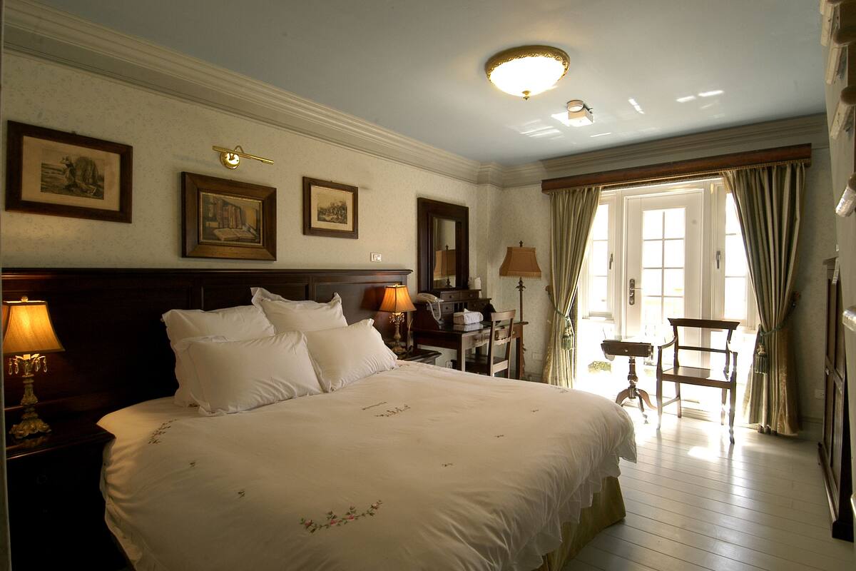 Une chambre d’hôtel avec un grand lit, deux tables de chevet avec des lampes, un bureau avec une chaise, un miroir et des tableaux encadrés accrochés au mur.
