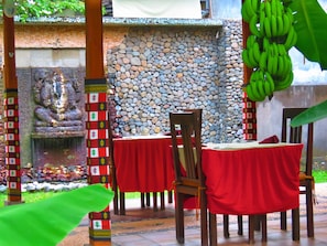 An outdoor dining area with stone walls, a Buddha statue, and tables with red tablecloths. - Suka Sari Cottages (Pemuteran)
