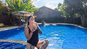 Una persona en traje de baño negro sentada junto a una piscina, con un cielo azul despejado y vegetación tropical al fondo.