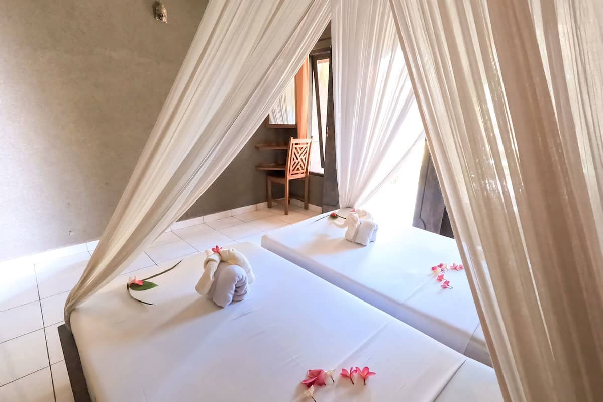 Ein Zimmer mit zwei Betten, jeweils mit Baldachin und Handtüchern. Ein Sessel und ein kleiner Tisch mit einer Blume sind ebenfalls vorhanden.