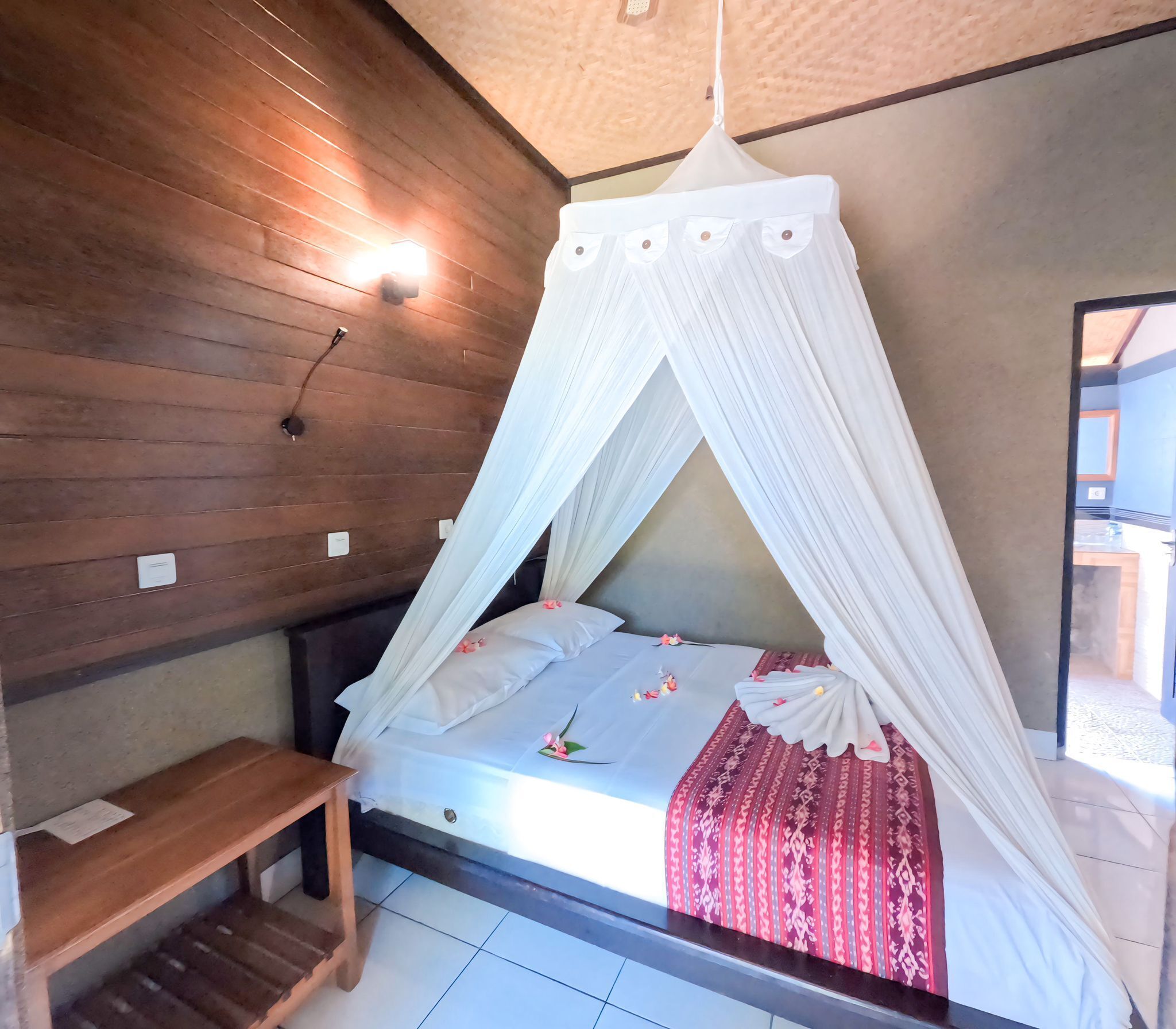 A bedroom with a wooden ceiling, a bed with a white canopy, a wooden bench, and a tiled floor.