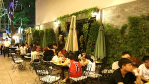 An outdoor cafe with tables and chairs, surrounded by greenery and decorative lights.