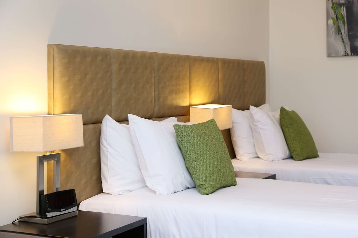 Habitación de hotel con dos camas, cada una con ropa blanca y almohadas verdes, una mesita de noche con una lámpara y un reloj digital.