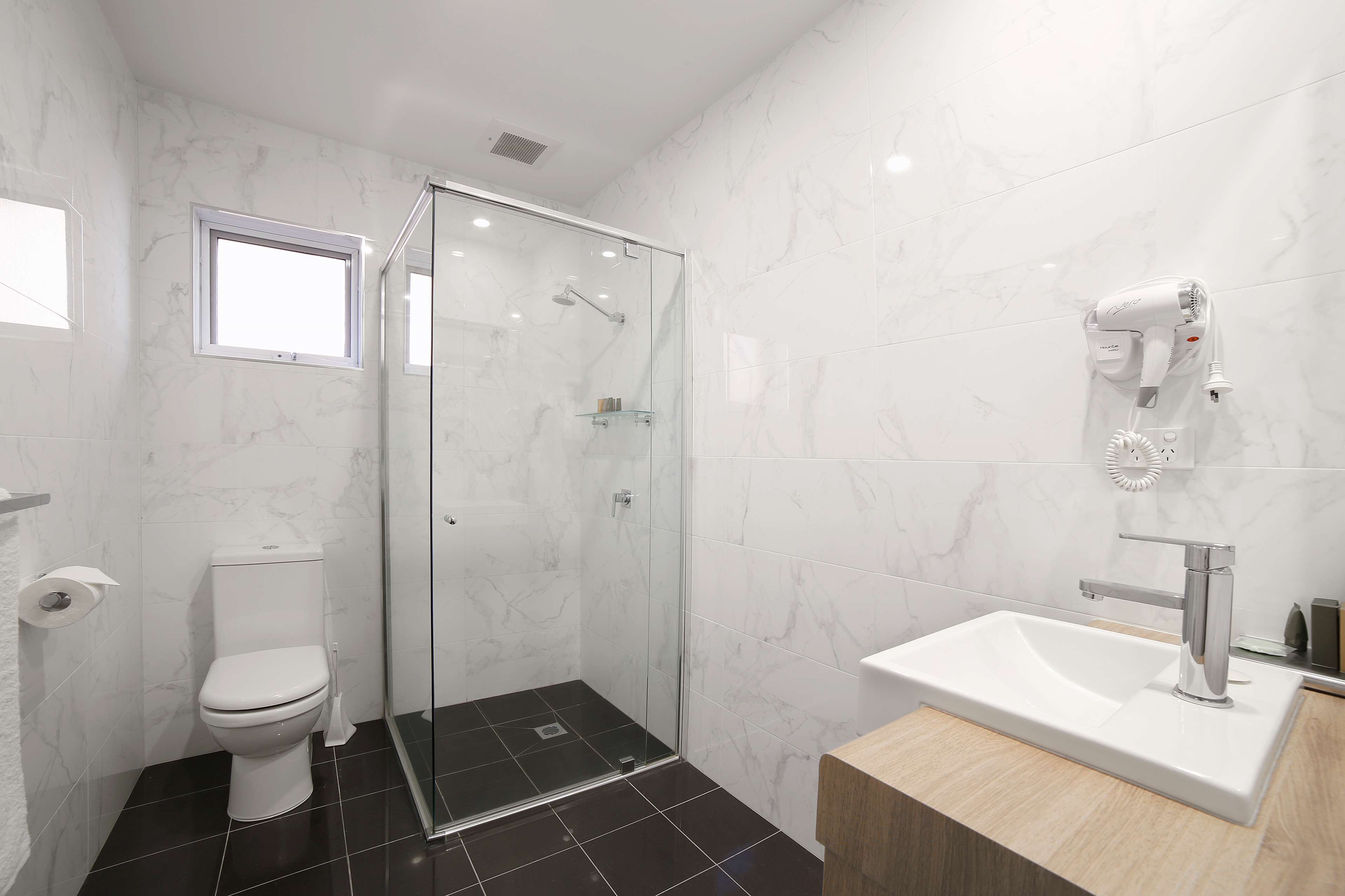 Ein modernes Badezimmer mit einer gläsernen Duschkabine, einem weißen Waschbecken mit Chromarmatur und einem an der Wand befestigten Föhn.