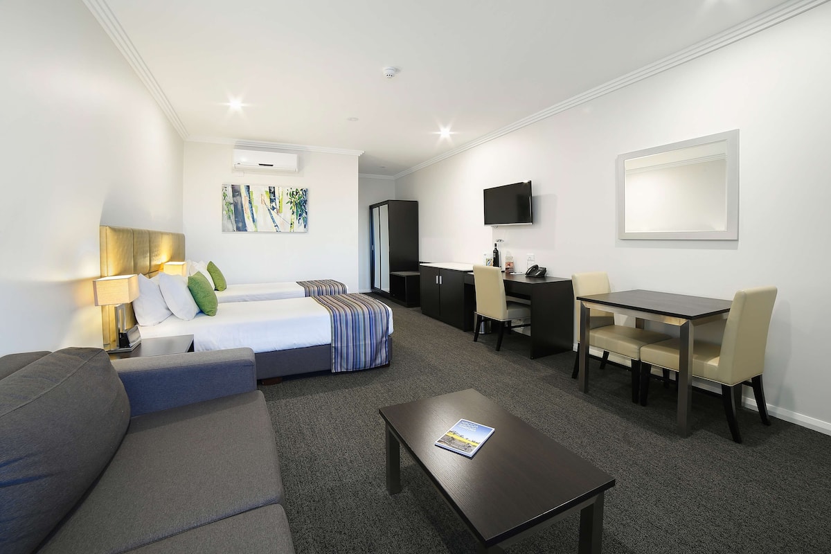 Habitación de hotel con cama, sofá, mesa de comedor con sillas, escritorio, televisor montado en la pared y piso alfombrado.