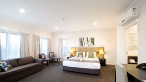 Habitación de hotel con una cama grande, un sofá, un escritorio y una silla. Cuenta con una ventana con cortinas, aire acondicionado instalado en la pared y un cuadro.