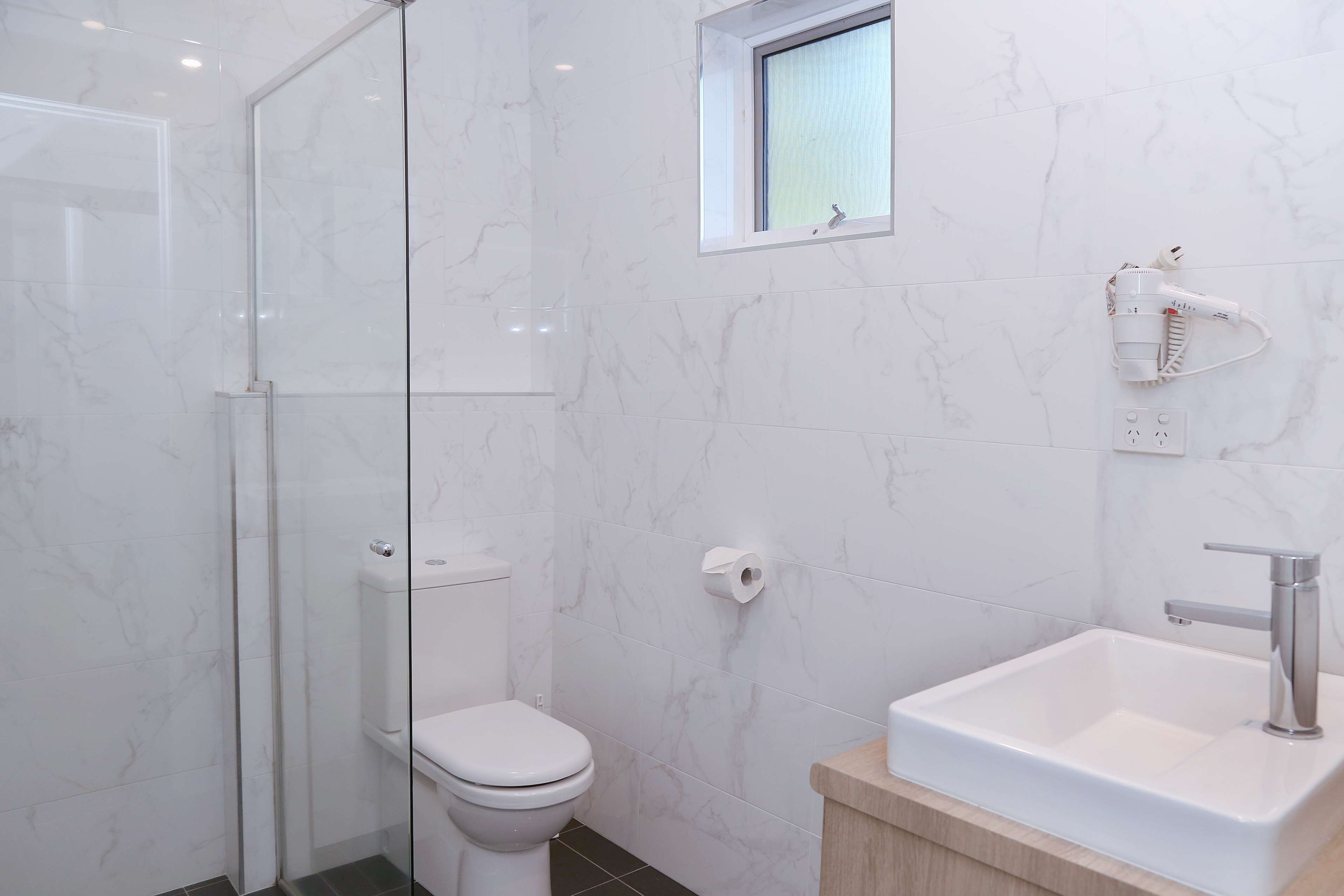 A modern bathroom with a glass shower enclosure, a toilet, a small window, and a sink with a mirror.