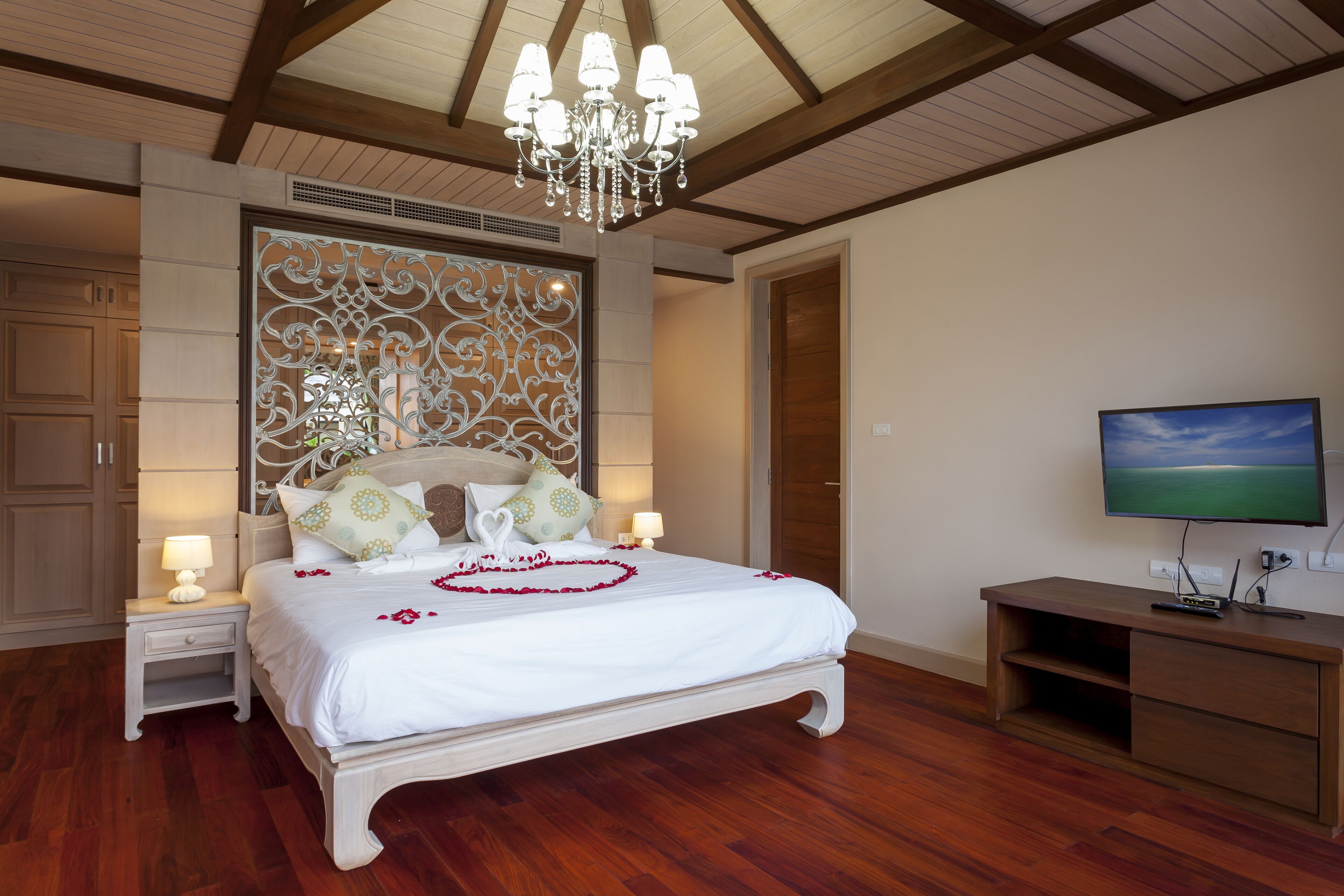 Un dormitorio amplio con una cama grande, piso de madera, un candelabro, un televisor y un panel decorativo en la pared.