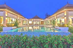 Una piscina con borde de azulejos, rodeada de vegetación y dos edificios con amplias ventanas, además de una estructura central.