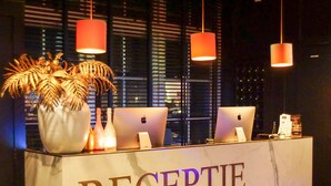 A reception desk with the word "RECEPTIE" on it, two laptops, a vase with palm leaves, and three hanging lamps. - Hotel Café Restaurant Dallinga (Sluiskil)