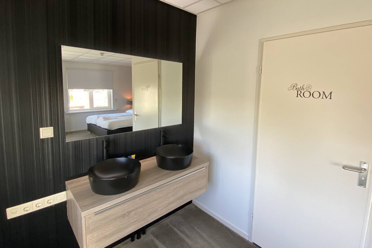 A bathroom with a large mirror, two black sinks, a wooden vanity, and a door labeled "Bath Room".