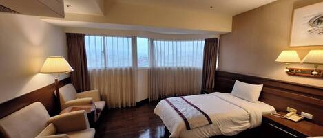 A hotel room with a bed, two chairs, a desk, a telephone, and a window with curtains.