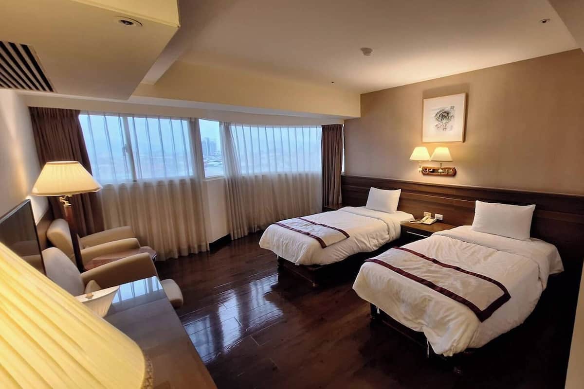 Habitación de hotel con dos camas, un escritorio, una silla, una lámpara y vistas a la ciudad a través de amplias ventanas.
