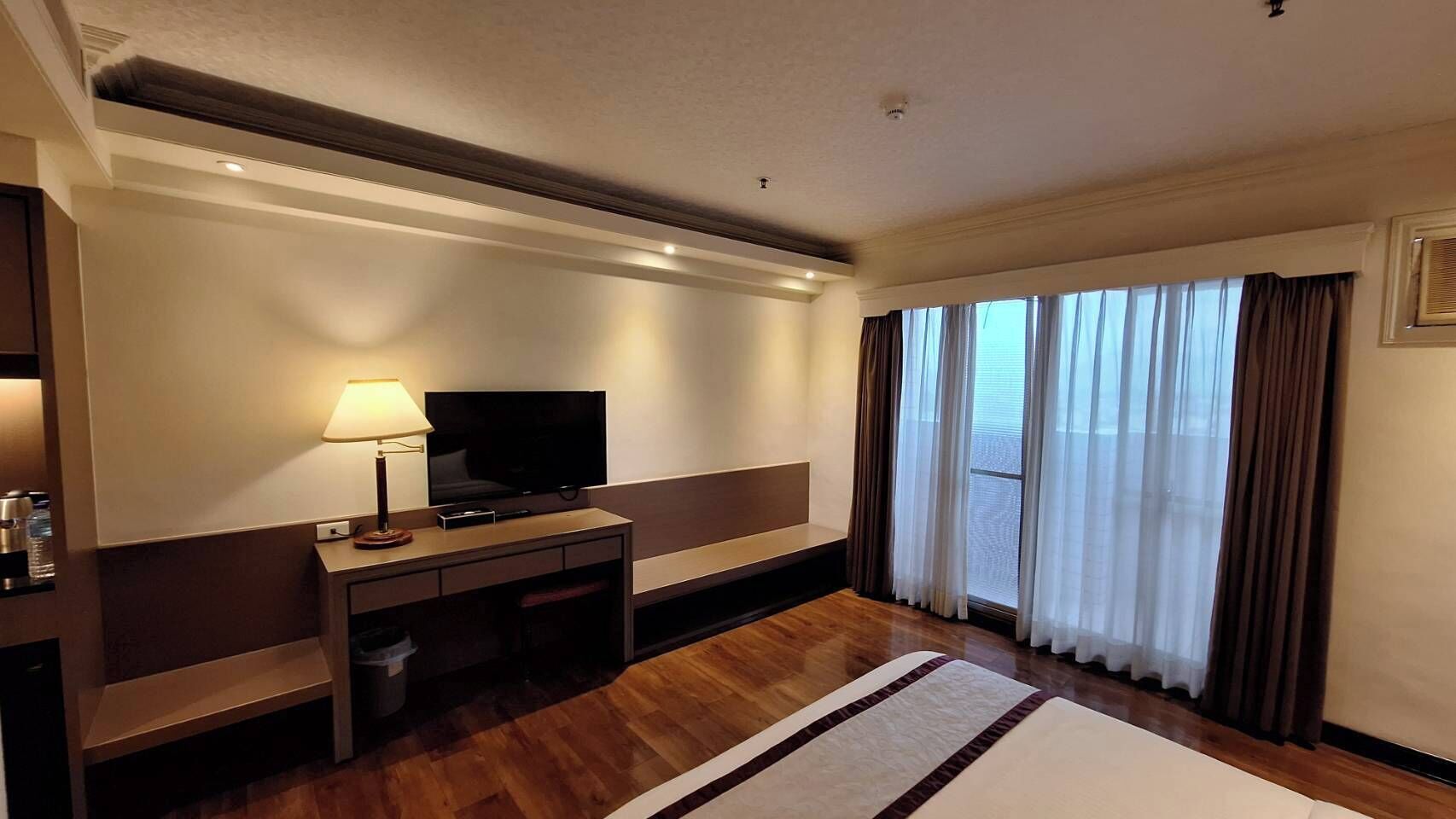 A hotel room with a bed, a desk with a TV, a lamp, and a window with curtains.