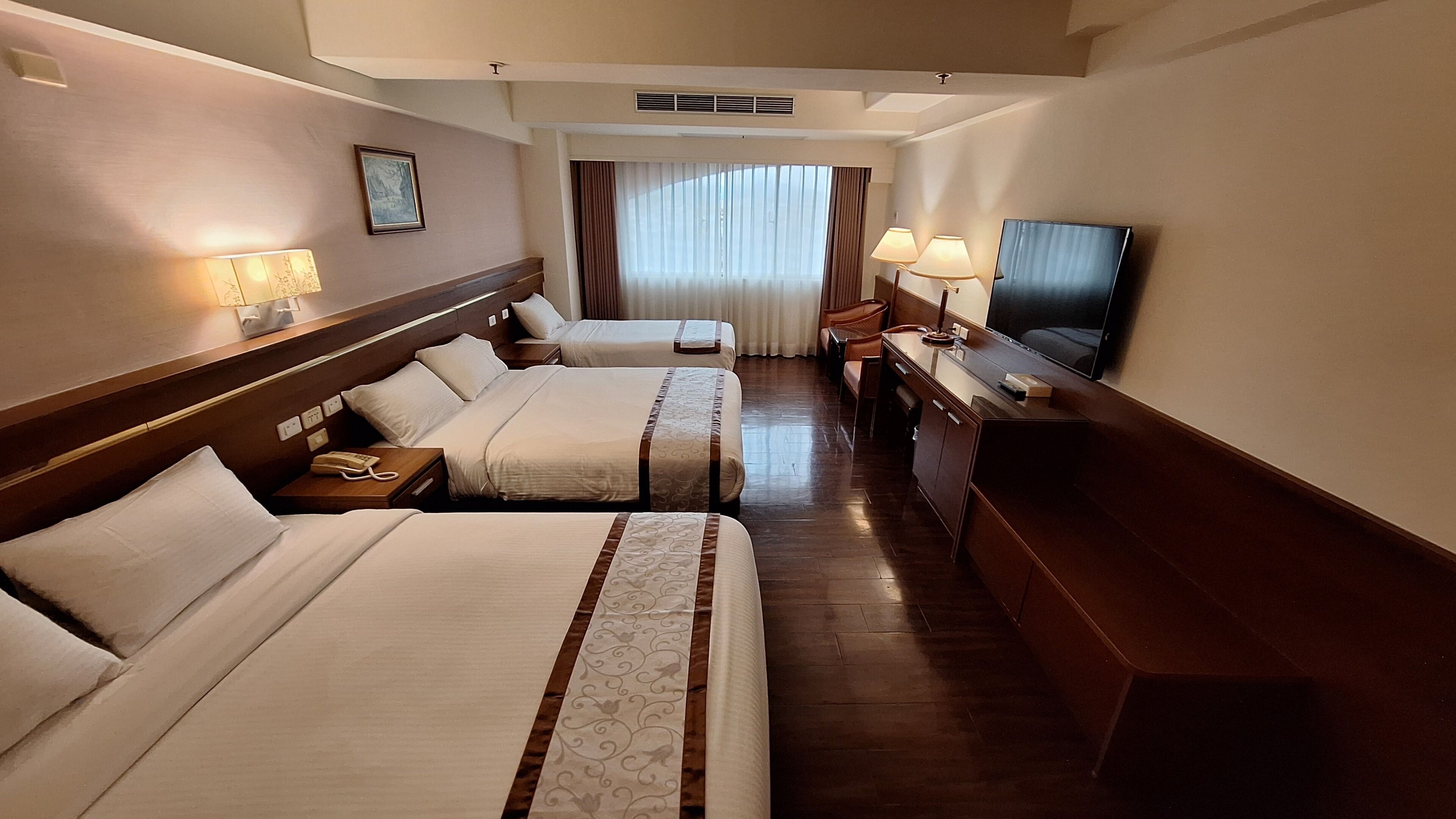 Ein Hotelzimmer mit zwei Betten, einem Schreibtisch, einem Fernseher und einem Fenster mit Vorhängen.