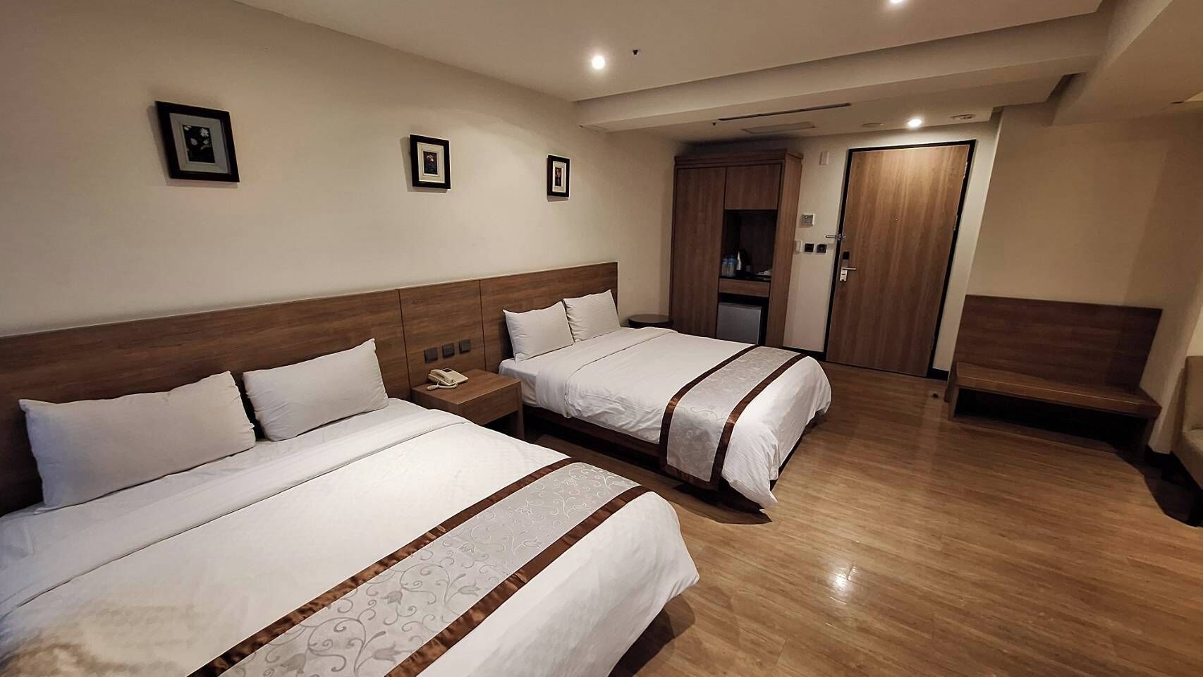 A hotel room with two beds, wooden headboards, a bedside table with a phone, and a wardrobe.