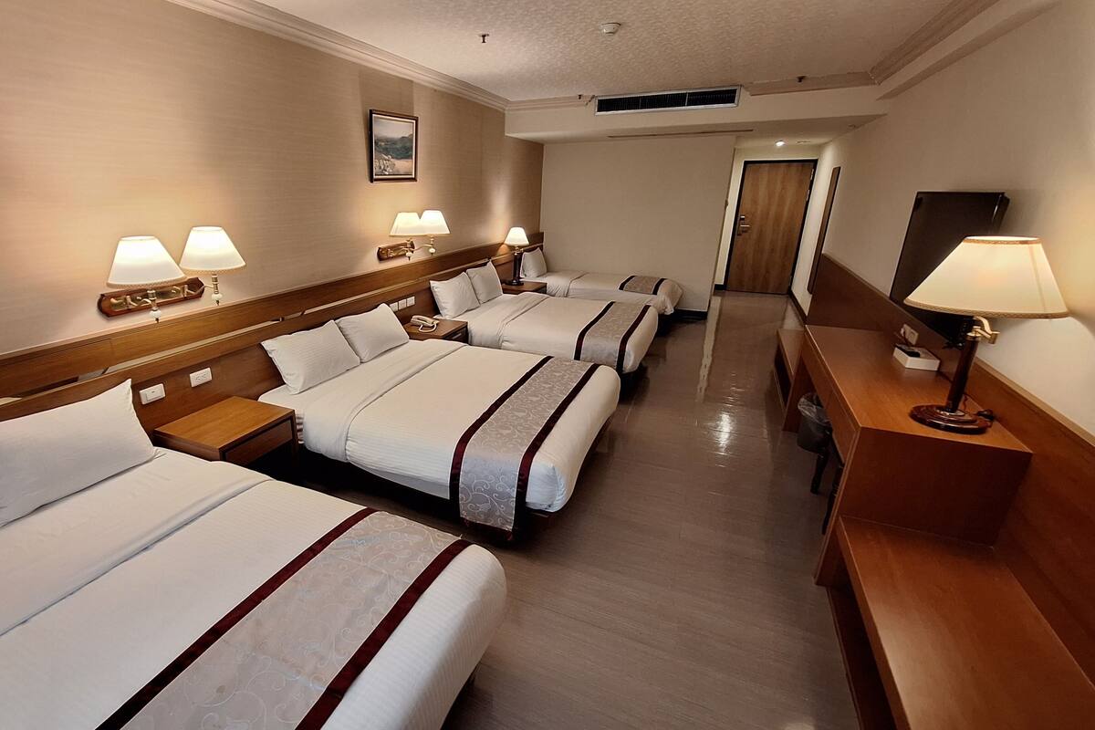 Habitación de hotel con cuatro camas dispuestas en fila, cabecera de madera, lámparas de noche, mesitas de noche de madera y un escritorio de madera con una lámpara.