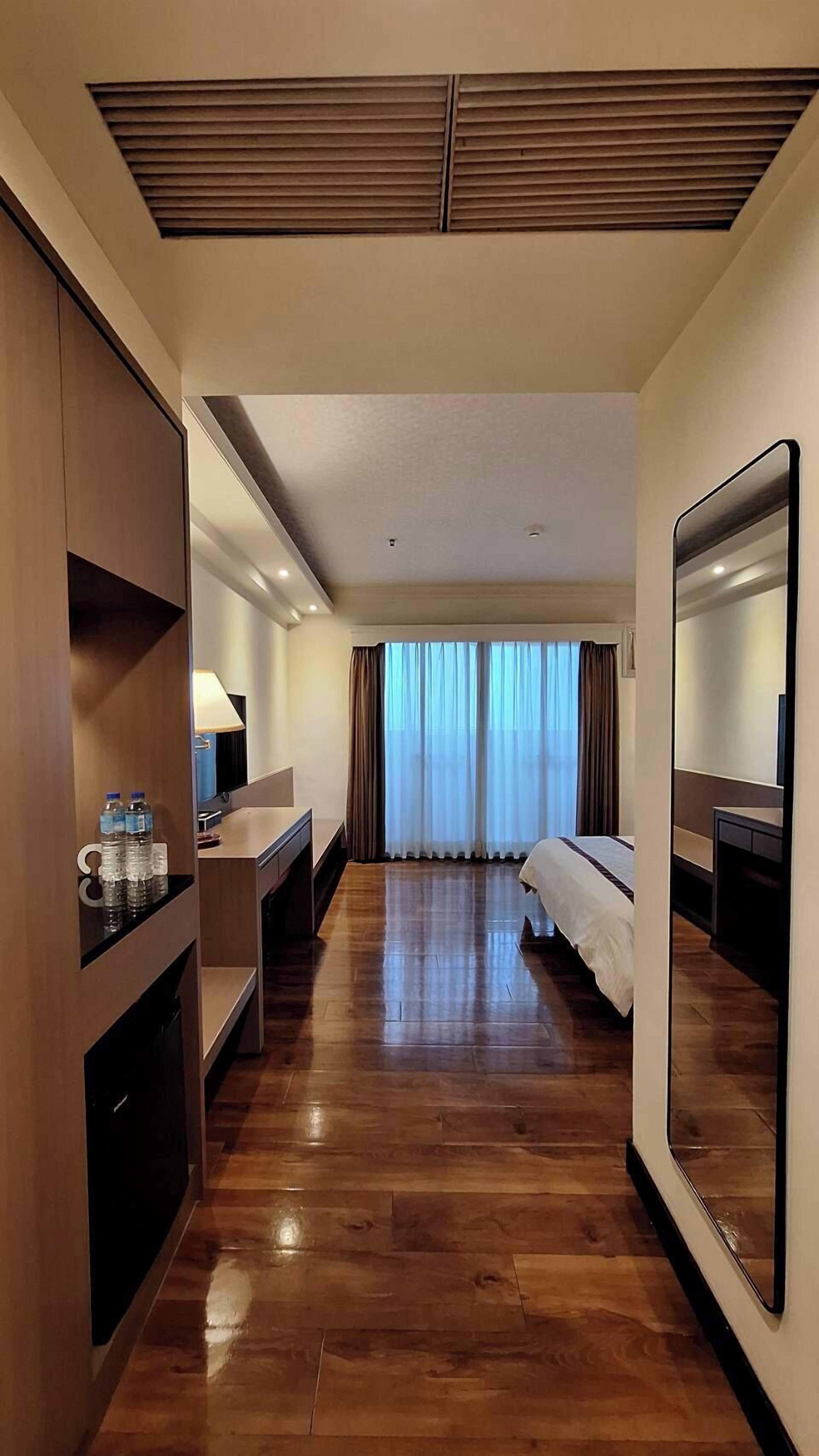 Una habitación de hotel con suelo de madera, cama, escritorio con silla, espejo y ventana con cortinas.