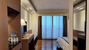 Una habitación de hotel con suelo de madera, cama, escritorio con silla, espejo y ventana con cortinas.