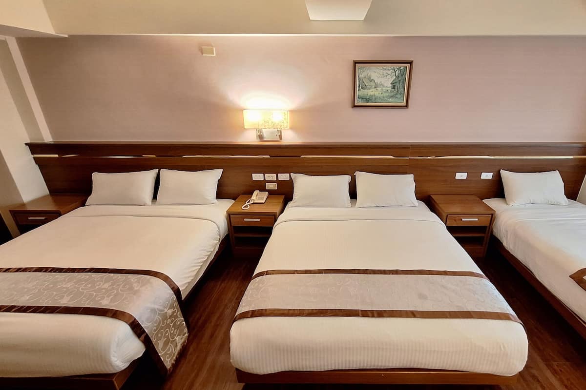 Habitación de hotel con tres camas individuales, un cabecero de madera, una mesita de noche con lámpara y un cuadro en la pared.