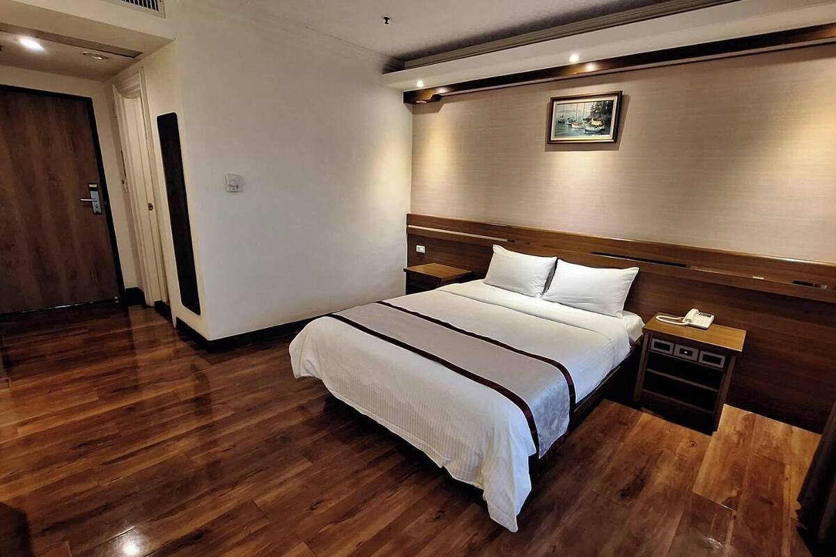 Habitación de hotel con piso de madera, cama con ropa blanca, mesita de noche y un cuadro en la pared.