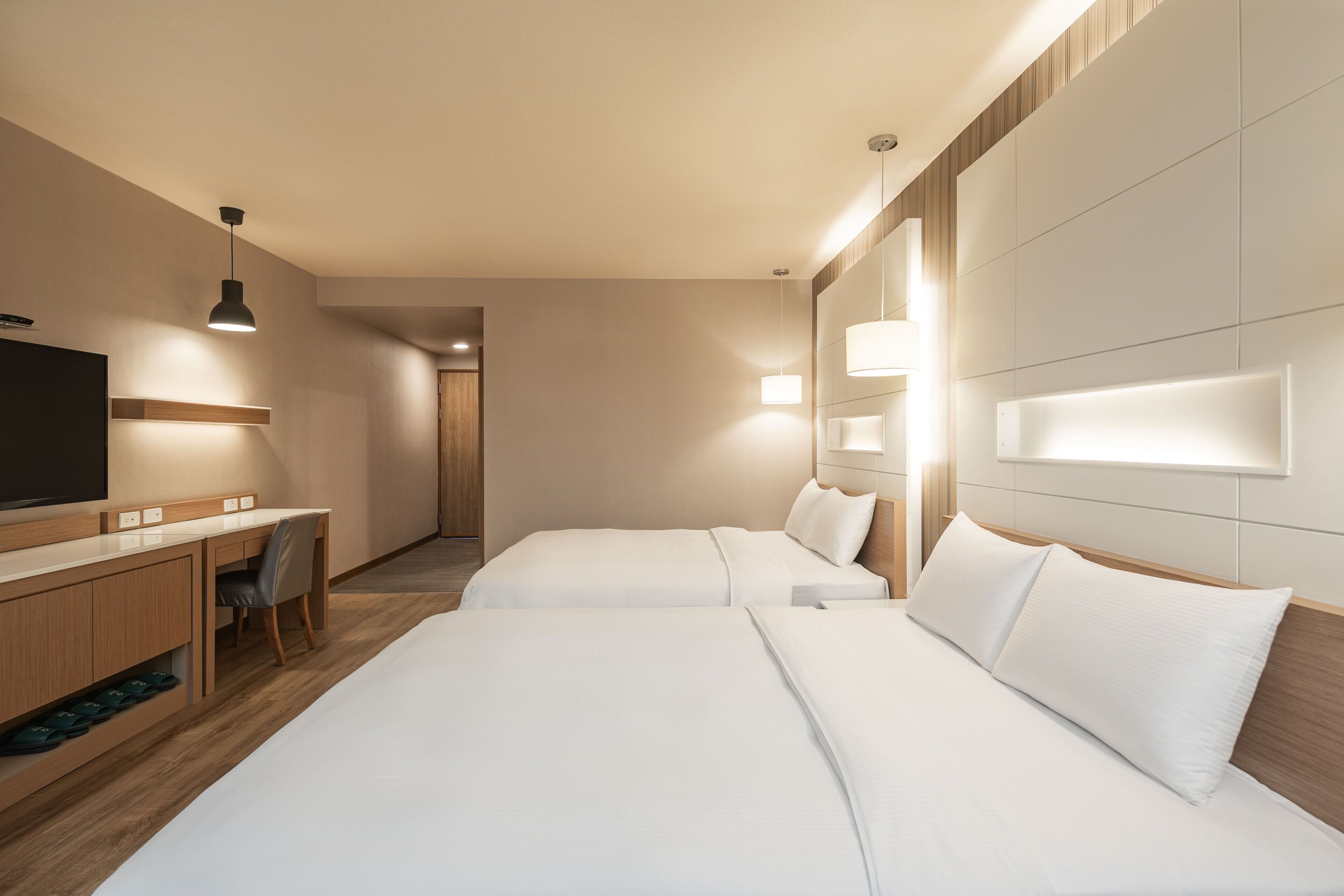A hotel room with two beds, a desk with a chair, a TV, and wall-mounted lights.