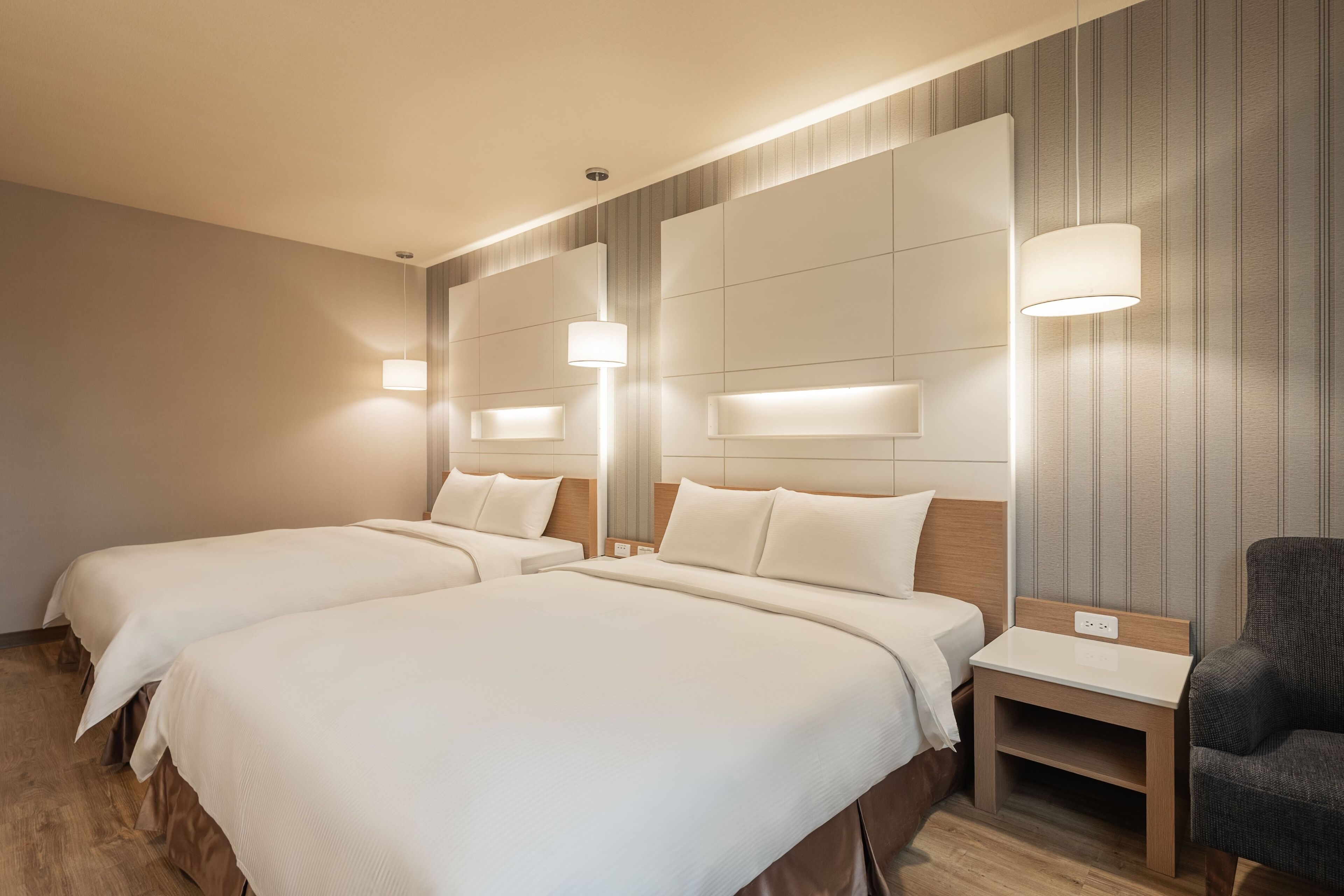 Habitación de hotel con dos camas, una mesita, una silla y una lámpara en la pared.