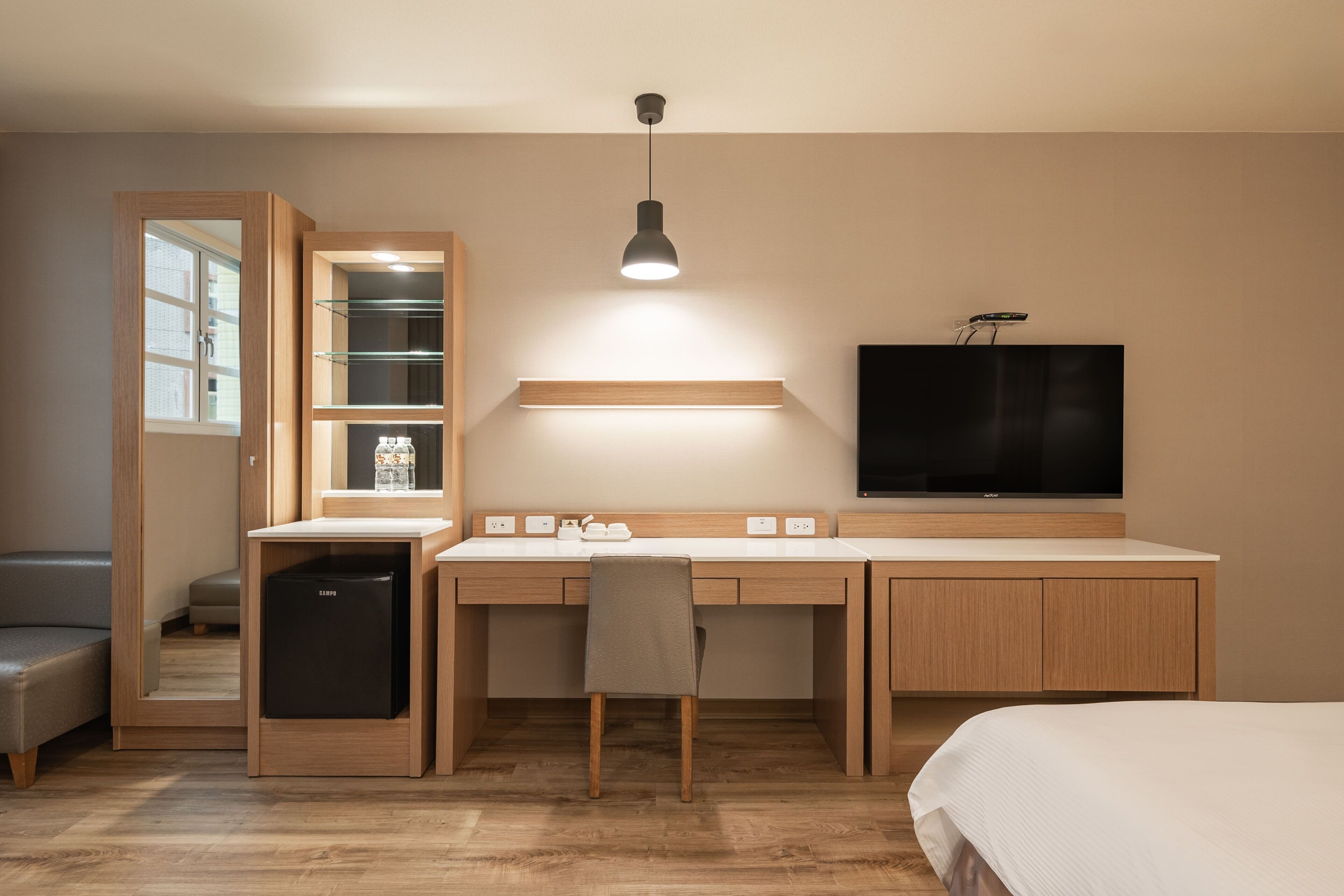 A compact hotel room with a wooden desk, a television, a bed, and a small kitchenette.