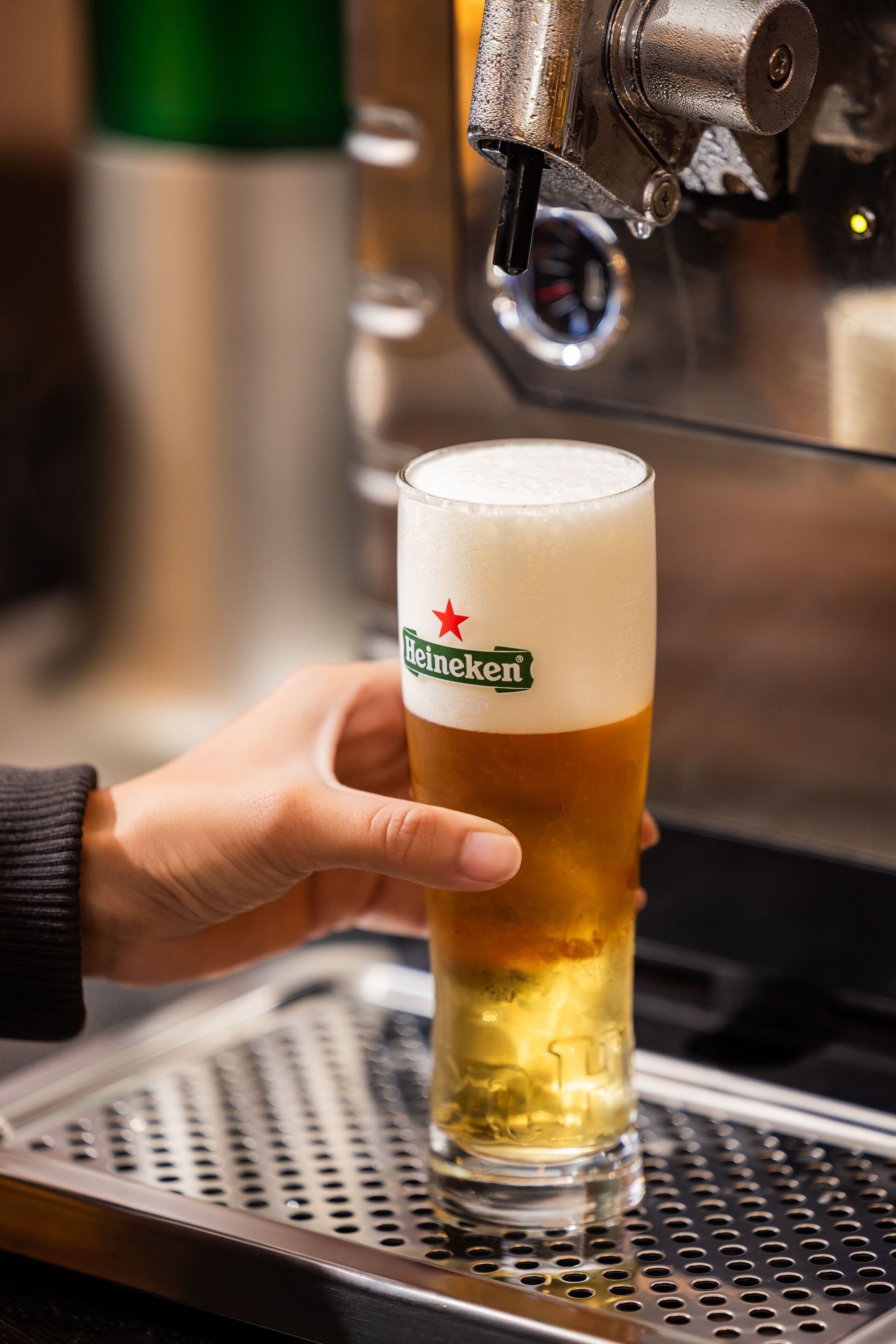 Una mano sosteniendo una cerveza Heineken servida en un vaso, frente a una llave de cerveza.