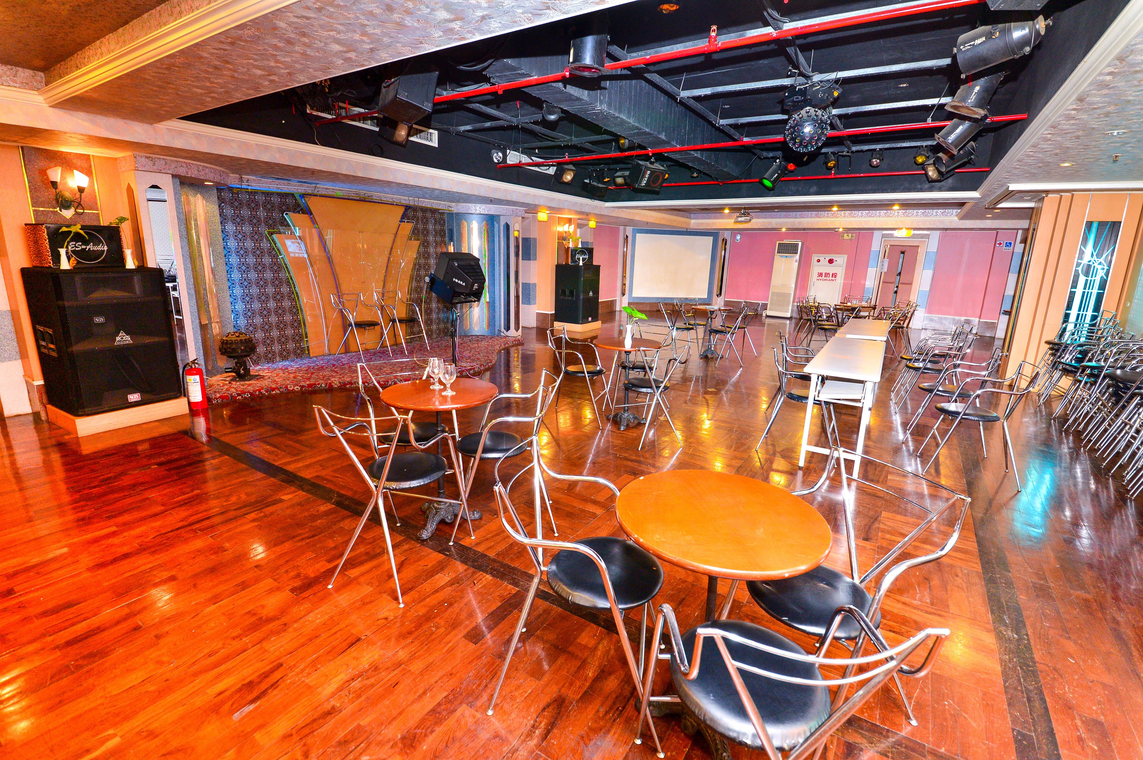 A spacious room with wooden flooring, multiple tables and chairs, and a stage area with sound equipment.