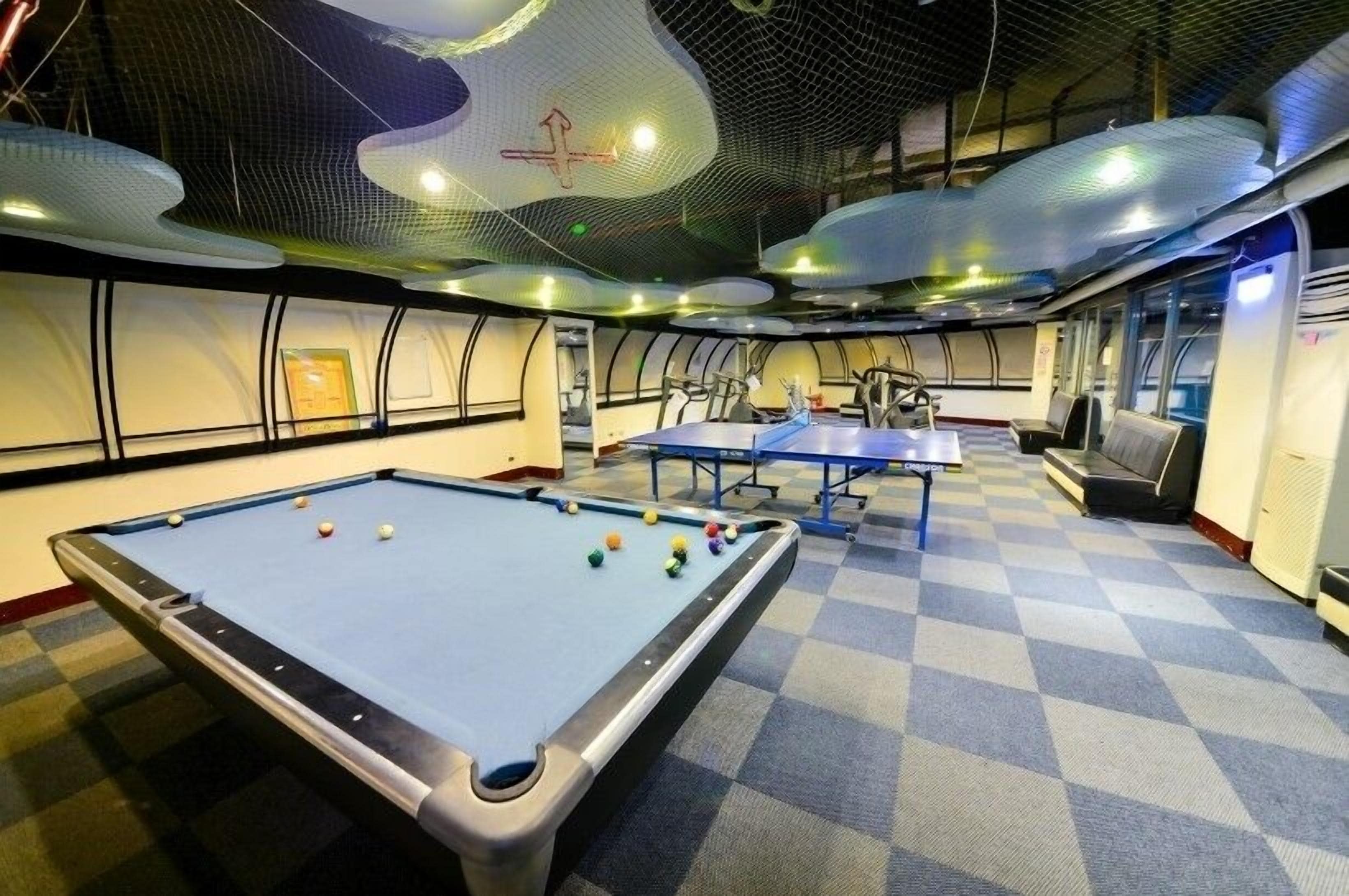 A pool table with balls, a ping pong table, and a billiards table in a room with checkered carpet and a high ceiling with a unique design.