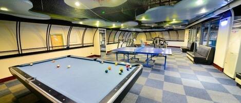 A pool table with balls, a ping pong table, and a billiards table in a room with checkered carpet and a high ceiling with a unique design.