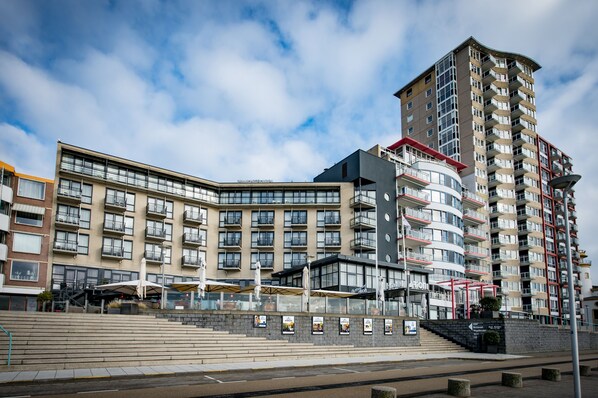 A modern multi-story hotel with a curved facade, outdoor seating area, and a prominent staircase leading to the entrance.