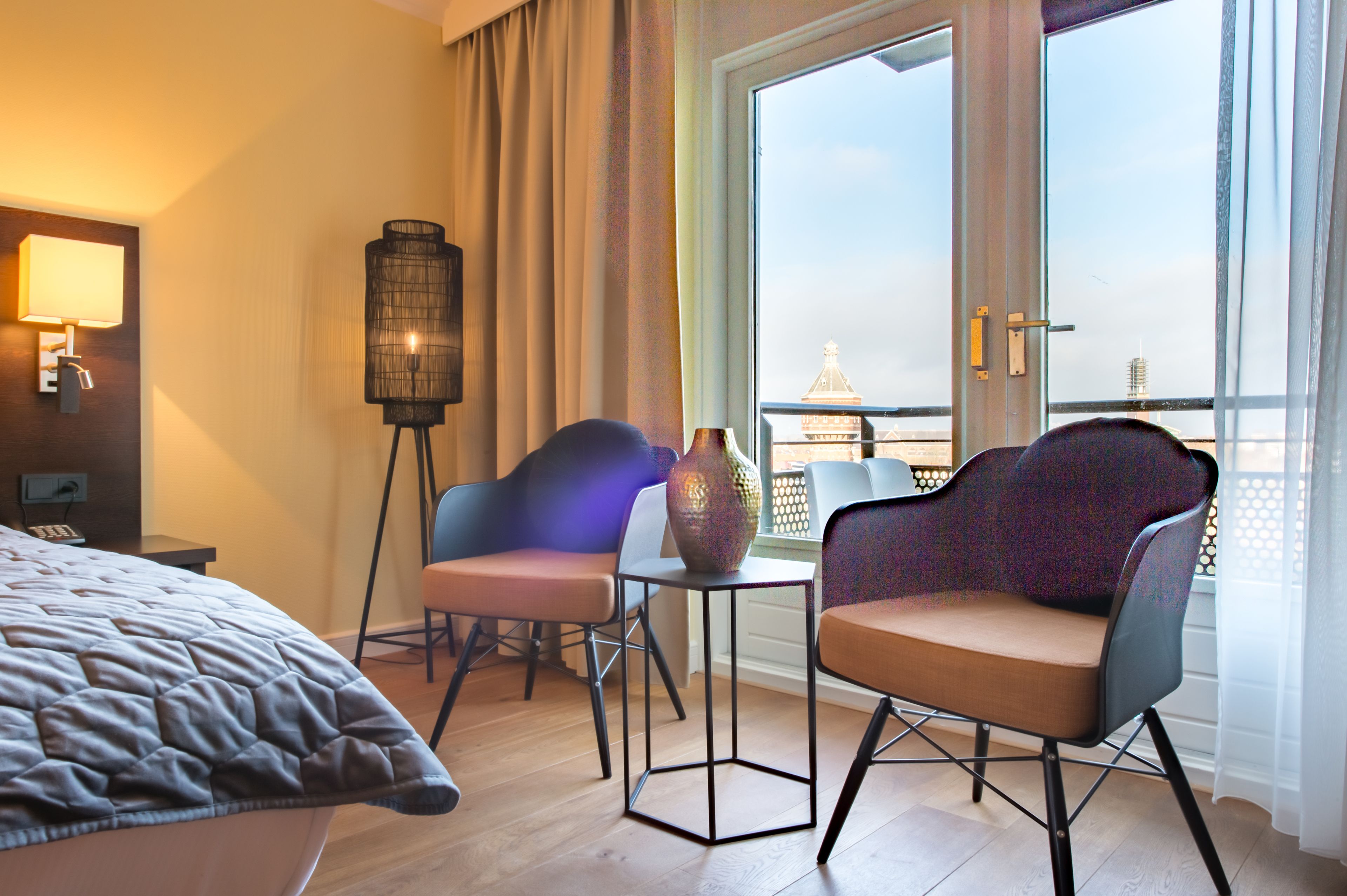 A hotel room with a bed, two chairs, a small table, a floor lamp, and a view of a cityscape through a large window.