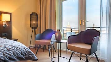 A hotel room with a bed, two chairs, a small table, a floor lamp, and a view of a cityscape through a large window.
