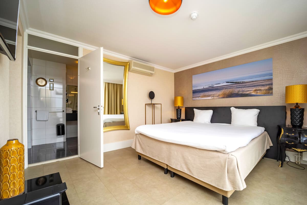 Habitación de hotel con una cama grande, un cuadro de playa, una mesita de noche con lámpara y un baño visible a través de una puerta abierta.