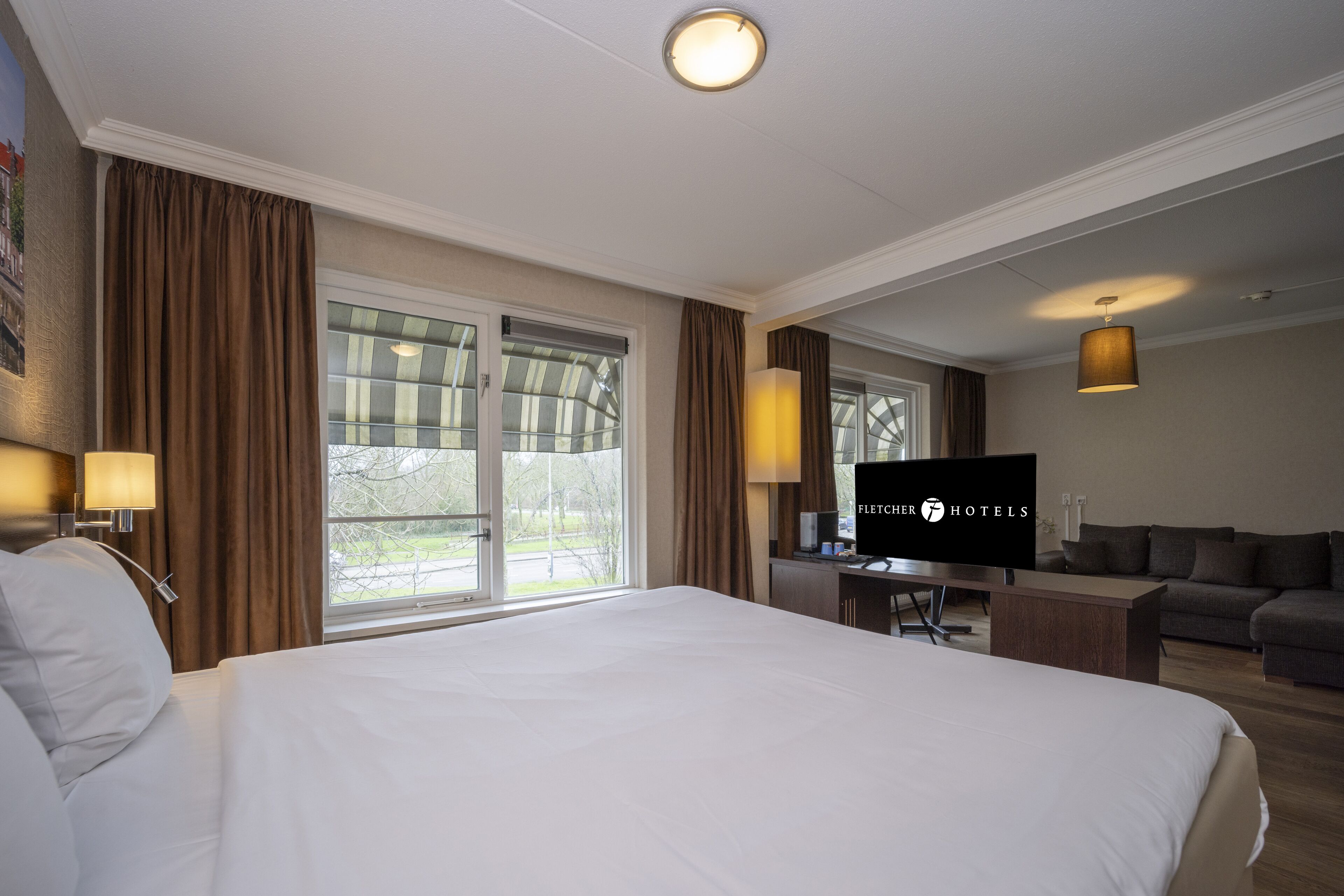 Una habitación de hotel con una cama grande, un escritorio con televisor, un sofá y una ventana con cortinas.