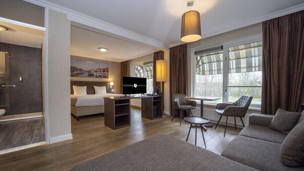 A hotel room with a sofa, a dining table with chairs, a TV, and a large window with curtains. - Fletcher Hotel-Restaurant Arneville-Middelburg (Middelburg)