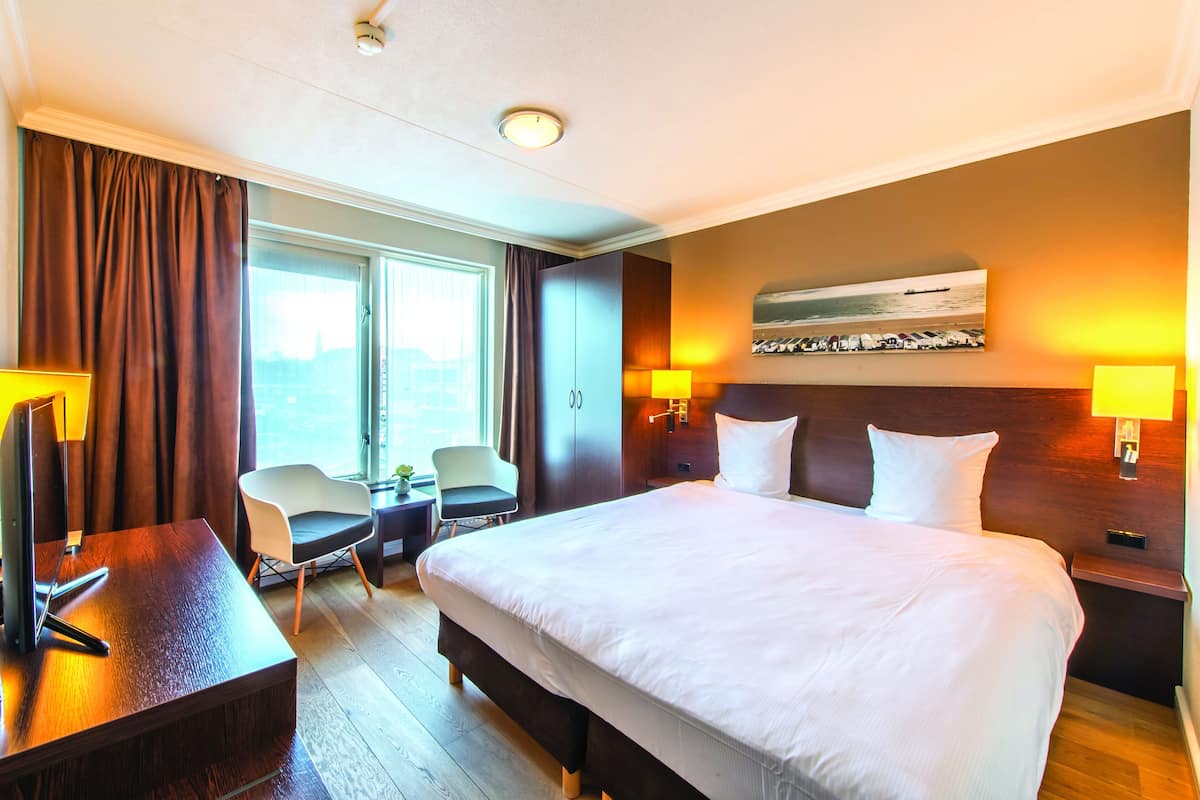 A hotel room with a large bed, two bedside tables, a desk with a chair, a small table with a chair, and a window with curtains.